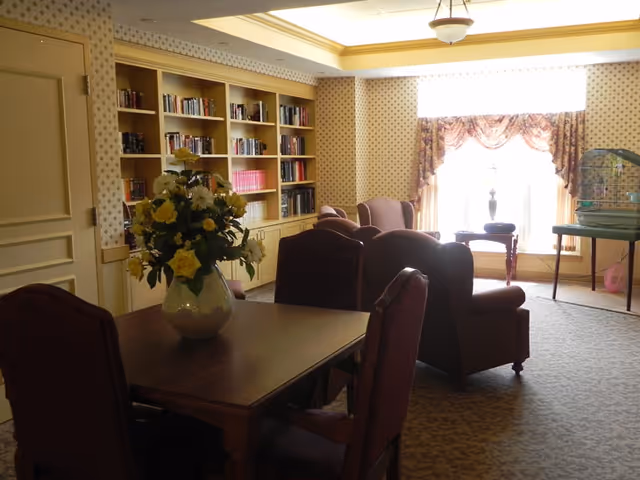 A cozy room with a wooden table and chairs in the foreground, a vase with yellow and white flowers on the table, several armchairs arranged near a large window with floral curtains, built-in bookshelves filled with books along one wall, and a birdcage on a stand near the window.
