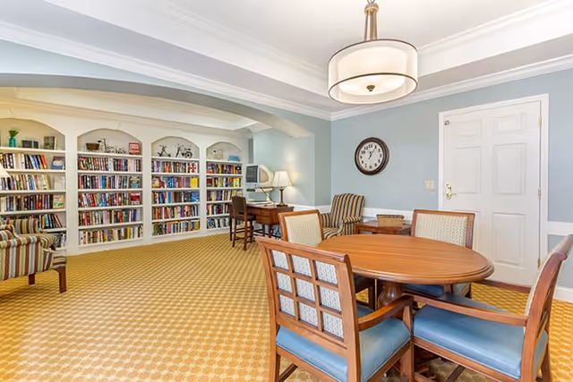 A bright communal interior room with built-in bookshelves, a round wooden table and chairs, armchairs, and a small desk under overhead lighting.