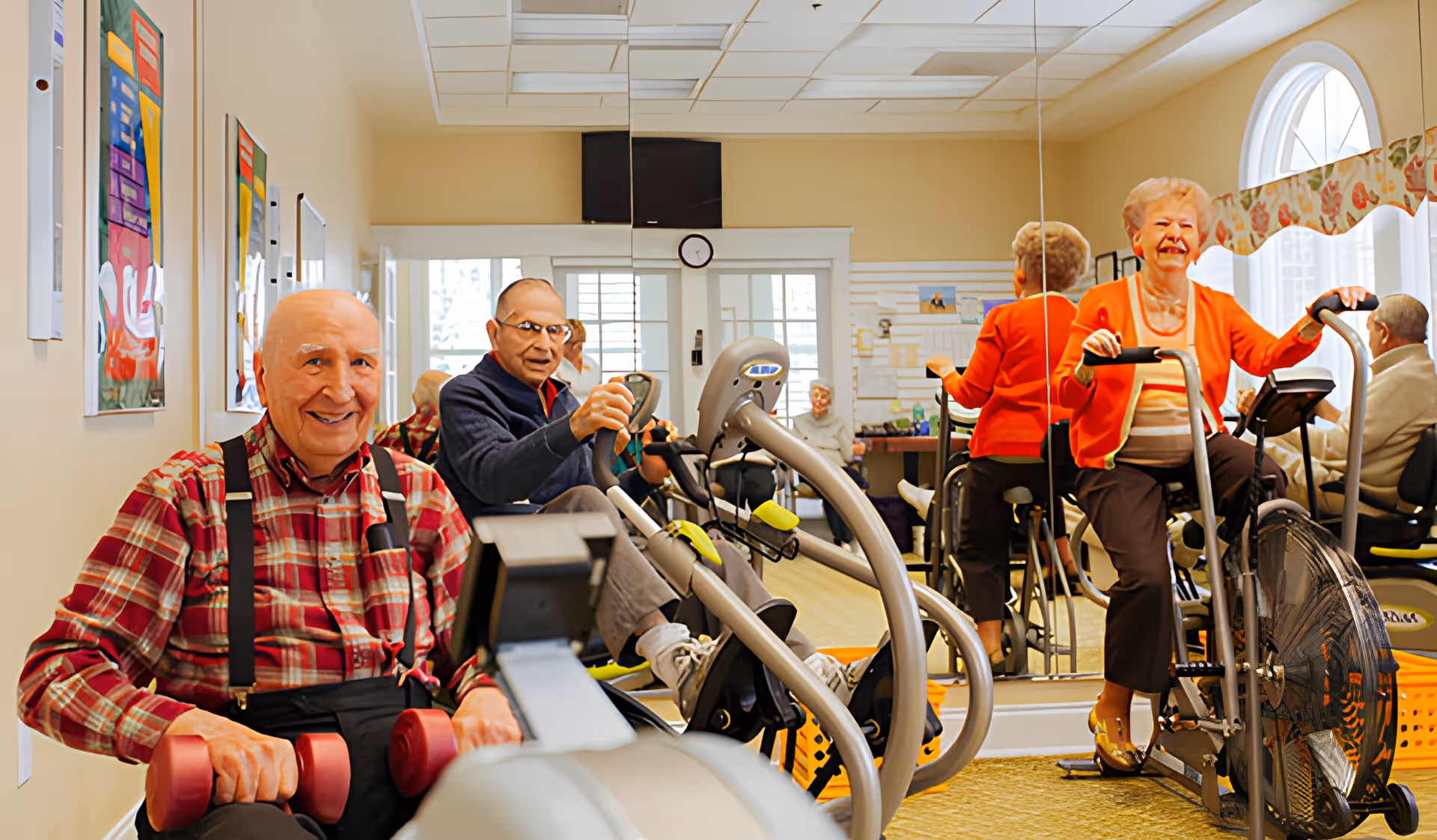 A group of elderly people exercising in a fitness room with exercise bikes and hand weights. The room has beige walls, a large mirror, colorful artwork on the walls, and a window with a floral valance. The seniors appear engaged and happy while using the exercise equipment.