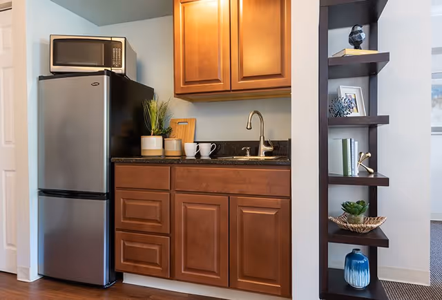 A small kitchen area featuring a stainless steel refrigerator with a microwave on top, wooden cabinets above and below a black countertop with a sink and faucet. On the countertop are two white mugs, a cutting board, and two small potted plants. To the right is a dark wooden shelving unit with decorative items including books, a small sculpture, and a blue vase.