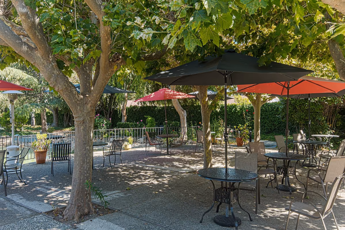 Shaded outdoor patio with tables, chairs, and colorful umbrellas beneath trees at a retirement community.