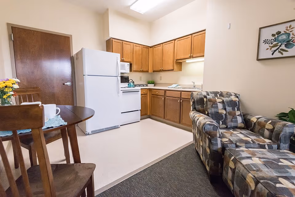 A small senior living facility kitchen and living area featuring wooden cabinets, a white refrigerator, stove, and microwave. There is a round wooden dining table with chairs and a vase of flowers on it. A patterned armchair and ottoman are positioned on a carpeted area next to the kitchen. A framed floral artwork hangs on the wall.