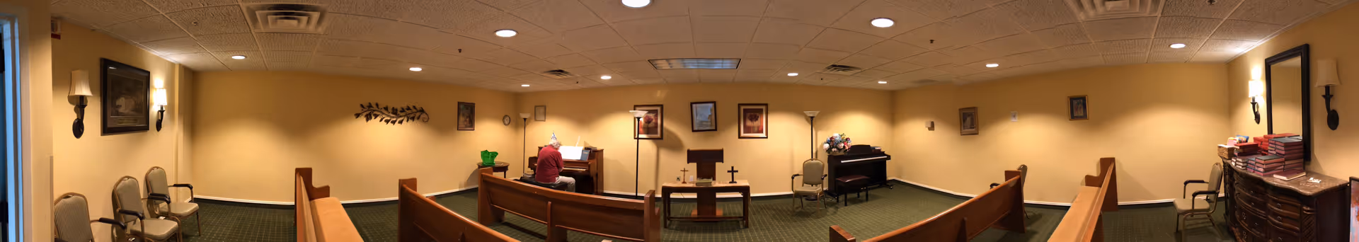 Interior of a small chapel or worship room with wooden pews arranged facing a podium and a piano. A person is seated at the piano playing music. The room has beige walls, green carpet, framed pictures on the walls, and soft lighting from wall sconces and ceiling lights.