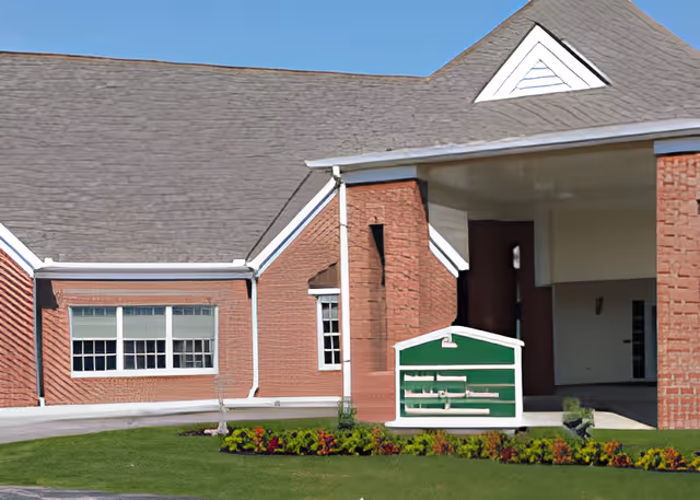 Exterior view of a brick building with a sloped roof and white trim. There is a covered entrance area and a small green and white mailbox or display box in front. The building is surrounded by a well-maintained lawn and flower beds under a clear blue sky.