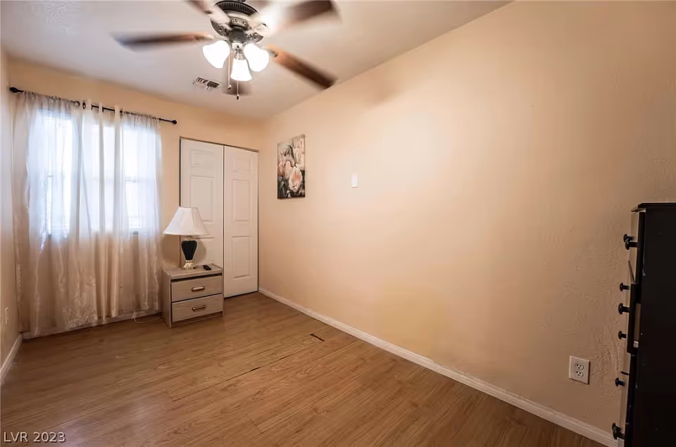 Small empty bedroom with wood floors, a ceiling fan, sheer-curtained window, nightstand with a lamp, and a closed closet.