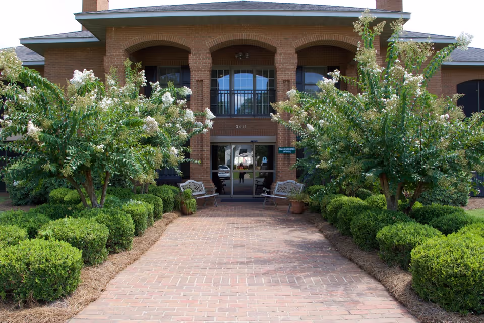 Front entrance of a brick building with a paved walkway lined by neatly trimmed bushes and flowering trees. Two benches and potted plants are placed near the entrance doors, which have a sign indicating the Sales Director Office.