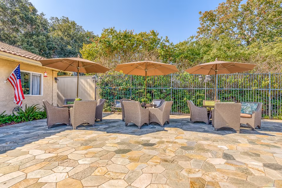 Outdoor patio area with stone tile flooring, featuring three sets of wicker chairs and tables, each shaded by a large beige umbrella. The patio is adjacent to a building with a window and an American flag hanging on the wall. Behind the patio is a black metal fence and dense green foliage under a clear blue sky.