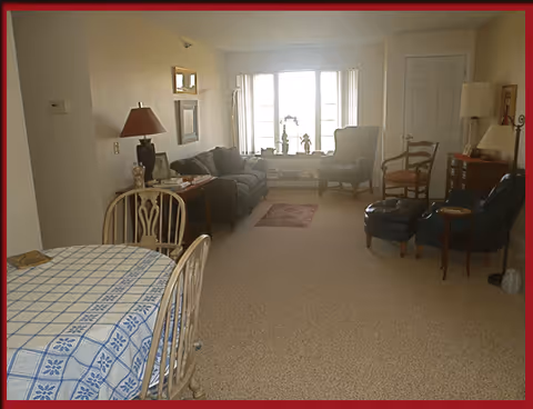 A cozy living room area with a beige carpet, a dark sofa, two armchairs, a wooden side table with a lamp, and a dining table with a blue and white checkered tablecloth and wooden chairs. There is a large window with white curtains letting in natural light, and various decorative items on the window sill and walls.
