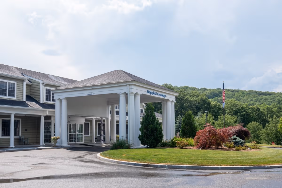 Exterior view of Benchmark at Ridgefield Crossings senior living facility showing the entrance with white columns and a covered driveway. There is a landscaped area with green grass, bushes, and an American flag on a flagpole. The building is two stories with beige siding and multiple windows.