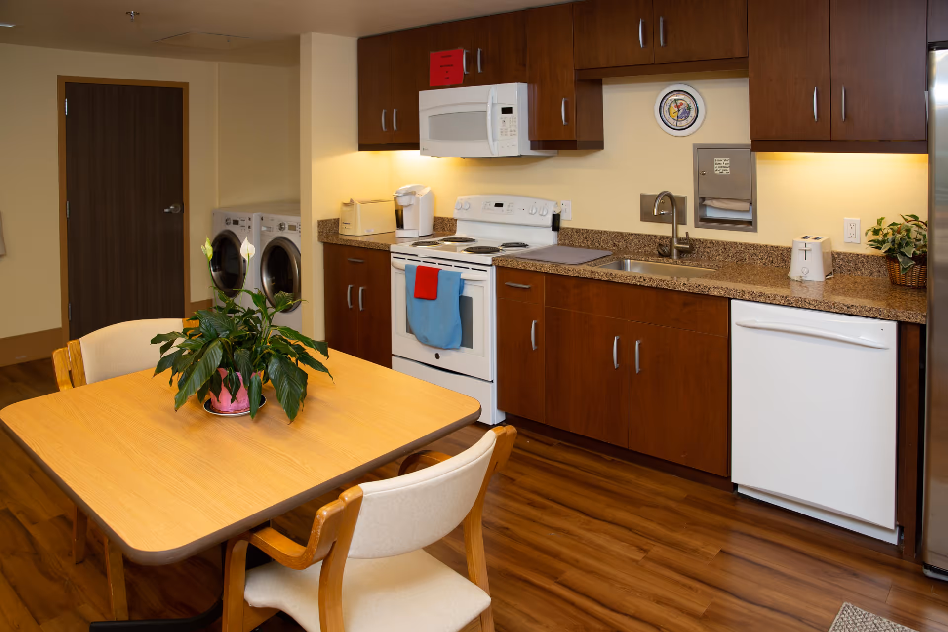 A kitchen area with wooden cabinets, a white stove with a red and blue towel hanging on the handle, a microwave above the stove, a toaster, a coffee maker, a sink, a dishwasher, and a refrigerator. In the foreground, there is a wooden table with four chairs and a potted plant on top. In the background, there is a door and a washer and dryer set.