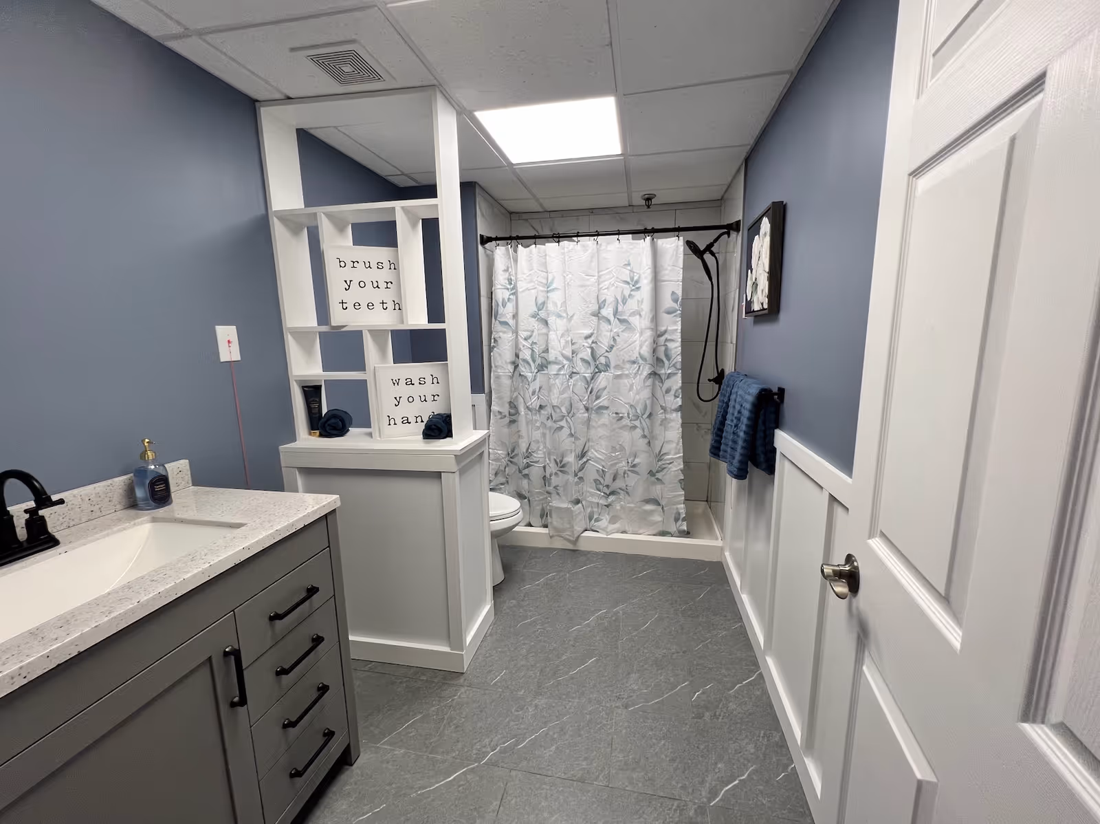 A clean and modern bathroom with gray and white tones. It features a gray vanity with a white countertop and black faucet on the left, a white shelving unit with signs that say 'brush your teeth' and 'wash your hands,' a toilet, and a shower with a white curtain decorated with blue leaves. Blue towels hang on a rack on the right wall, which is painted blue with white wainscoting. The floor is gray tile, and the ceiling has white panels with a bright light.