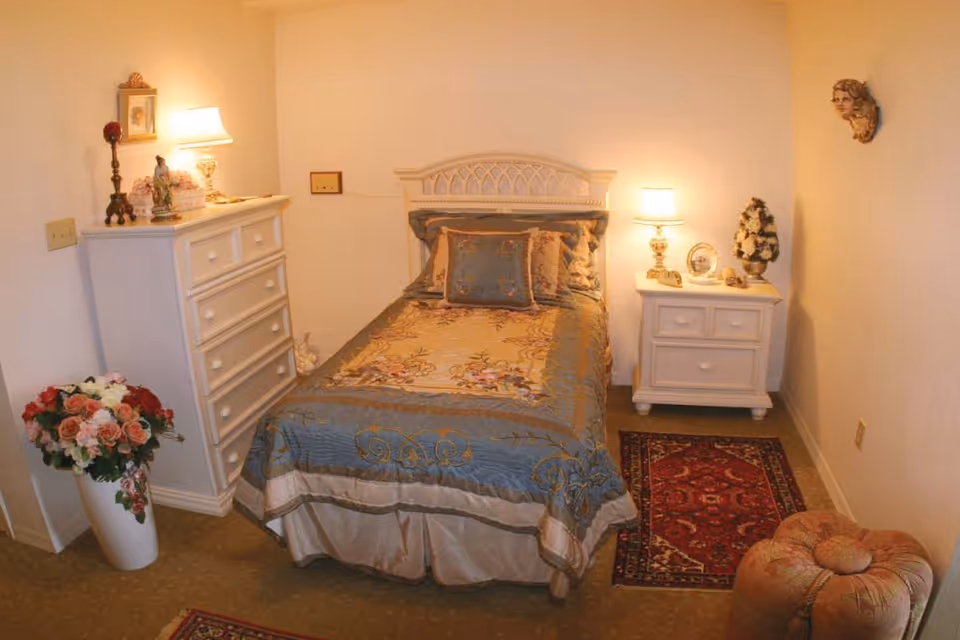 Small decorated bedroom with a single bed, floral bedding, a white dresser and nightstand with lamps, rugs, and a vase of flowers.