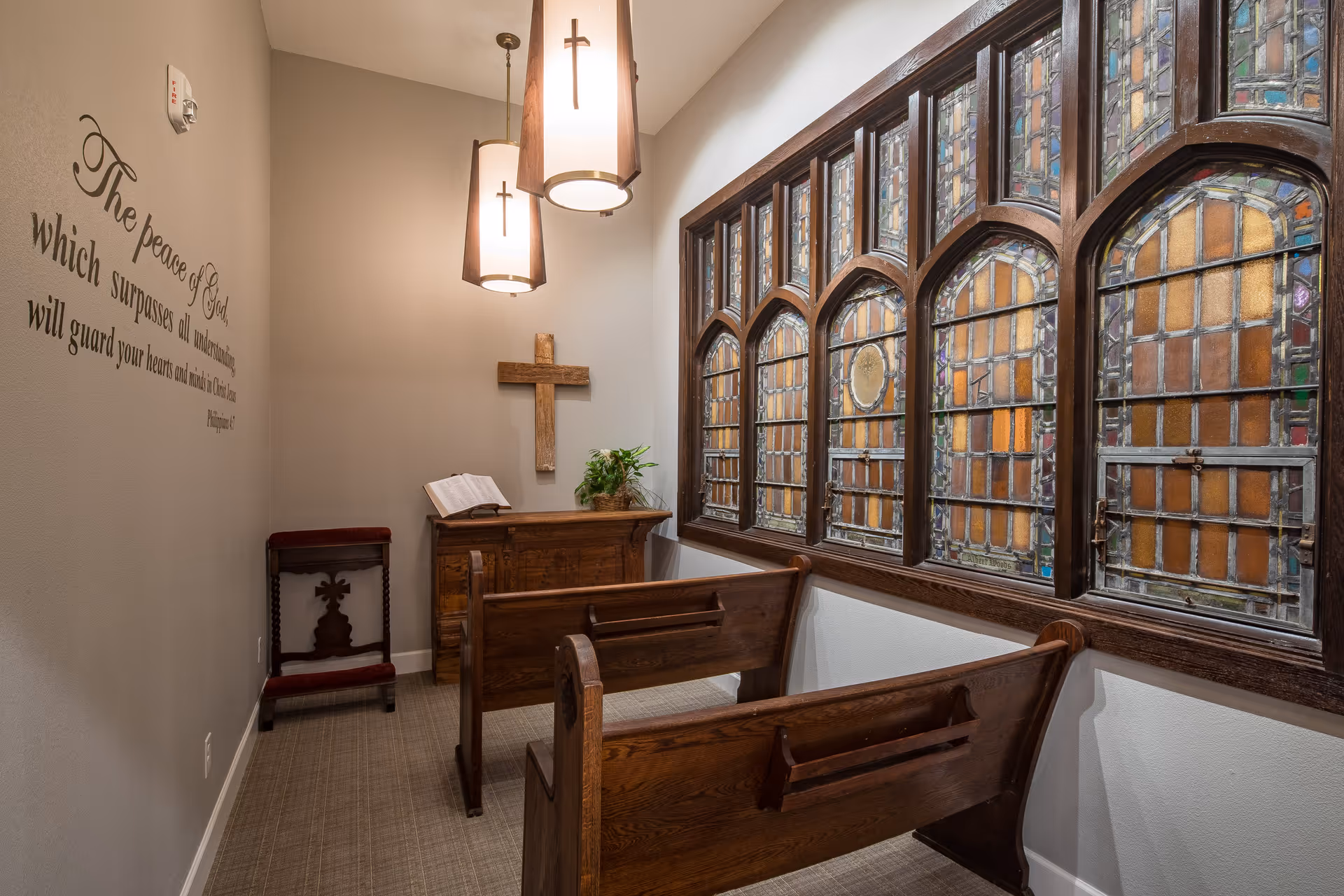 A small chapel room with wooden pews, a wooden lectern with an open book, a wooden cross on the wall, and stained glass windows. Two hanging lamps with cross designs illuminate the space. A religious quote is written on the left wall.