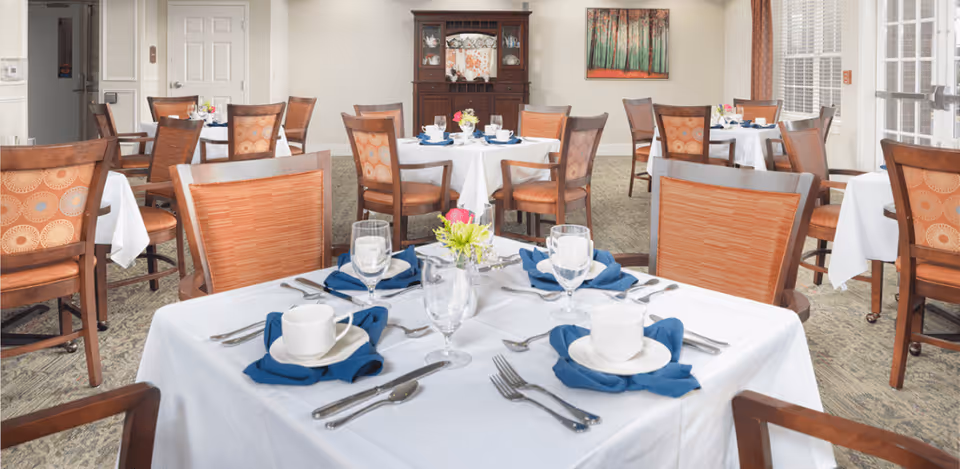 A dining room with several tables covered in white tablecloths, each set with white cups, plates, silverware, blue cloth napkins, and glasses. The room has wooden chairs with orange patterned cushions, a carpeted floor, a wooden cabinet against the far wall, and a painting of trees on the wall.