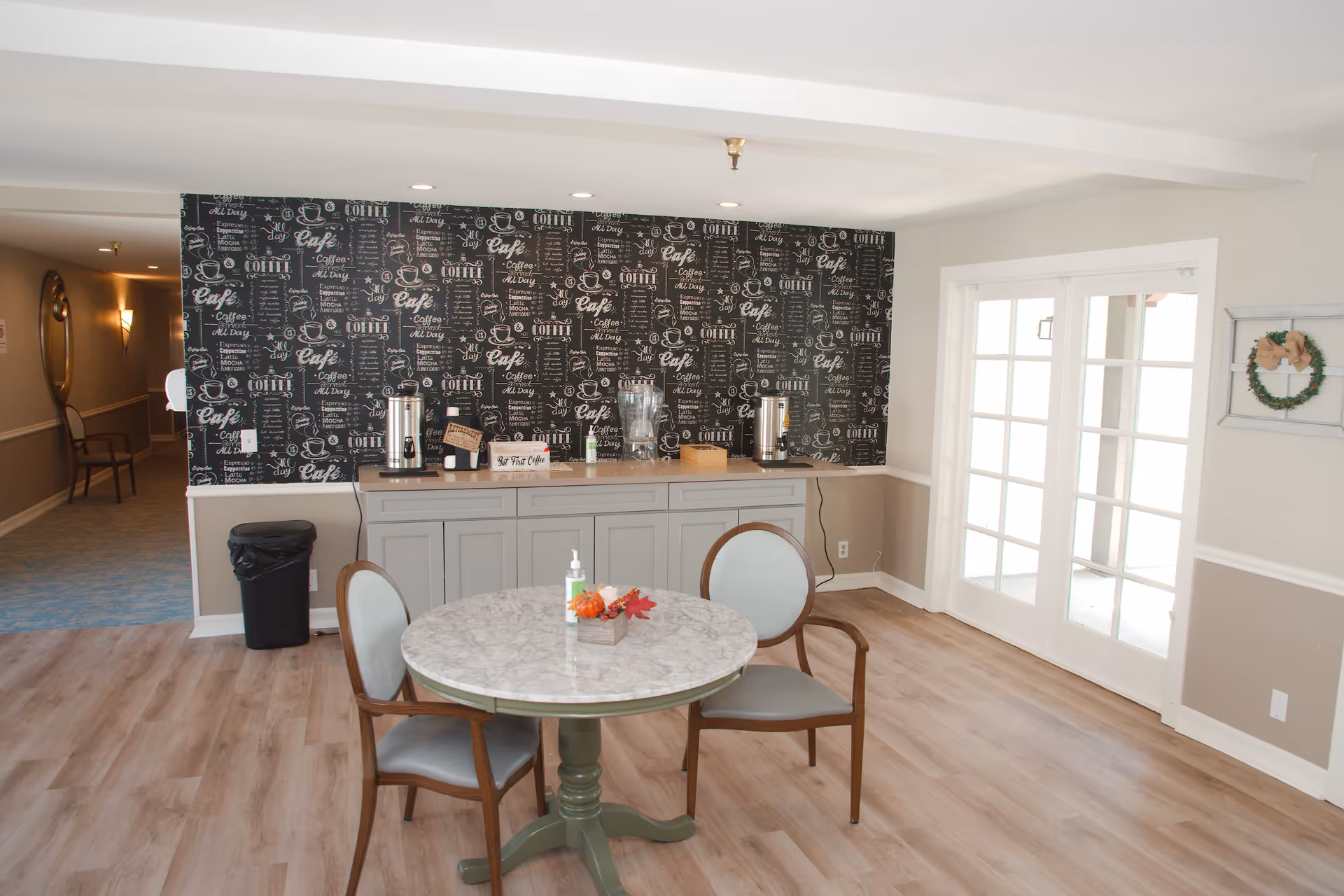 A cozy coffee station area in La Posada Assisted Living and Memory Care featuring a round marble table with two chairs, a countertop with coffee dispensers, water pitcher, and a decorative blackboard-style wallpaper with coffee-related words and illustrations. There is a hallway with a chair and a large round mirror on the left, and French doors on the right with a small wreath decoration on the wall.