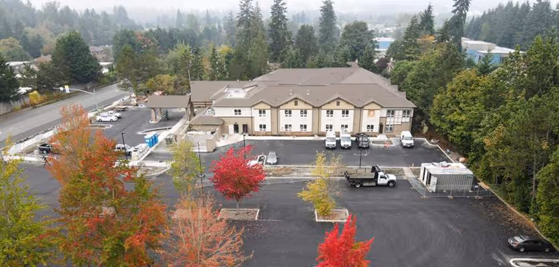 Aerial view of Silverado Bellevue Memory Care Community building surrounded by trees with autumn foliage. The building has a beige exterior with a brown roof and a parking lot with several vehicles parked. The area is bordered by dense green trees and a road is visible on the left side.