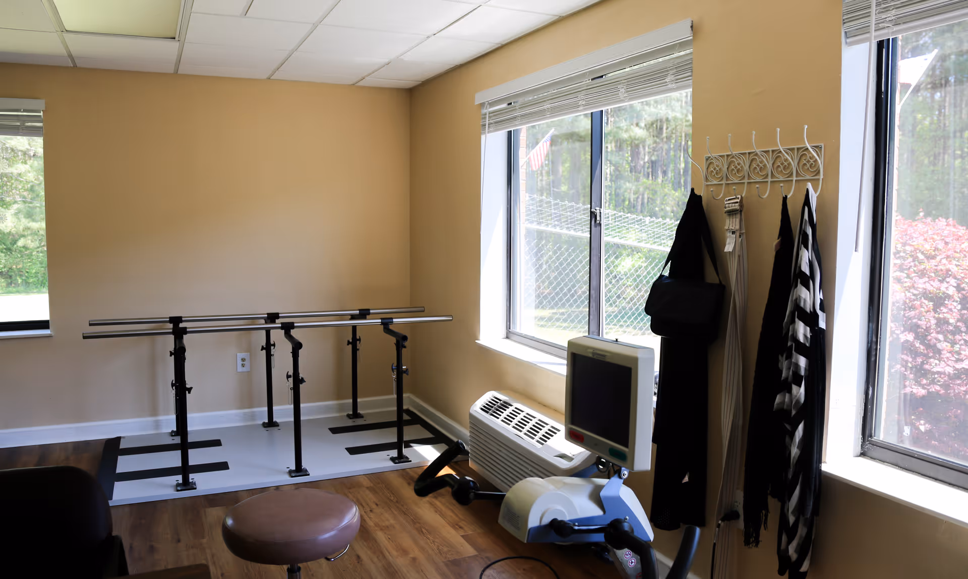A rehabilitation room with parallel bars for walking exercises, a stationary exercise bike with a monitor, a stool, and a coat rack with hanging bags and scarves. The room has beige walls, two large windows with blinds, and wood flooring.