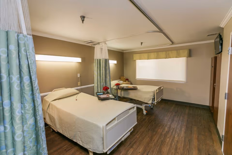 A room with two hospital-style beds separated by a curtain. Each bed has a beige bedspread and a small table with red flowers and magazines. The room has wooden flooring, beige walls, a large window with a closed blind, and a wall-mounted TV.
