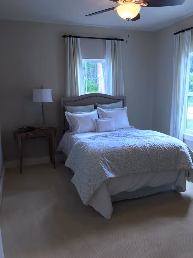 Bright, simply decorated bedroom with a made bed, bedside table and lamp beneath two windows and a ceiling fan.