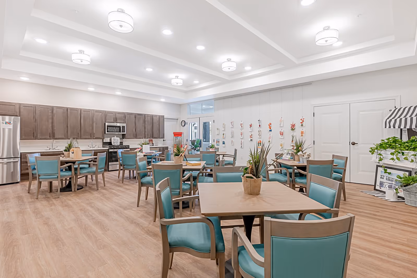 A bright and spacious dining area with multiple wooden tables and teal cushioned chairs. The room features a modern kitchen along one wall with stainless steel appliances, including a refrigerator, stove, and microwave. The walls are decorated with hanging floral artwork, and there are potted plants on the tables and a small plant stand on the right side. The ceiling has recessed lighting and several round light fixtures.