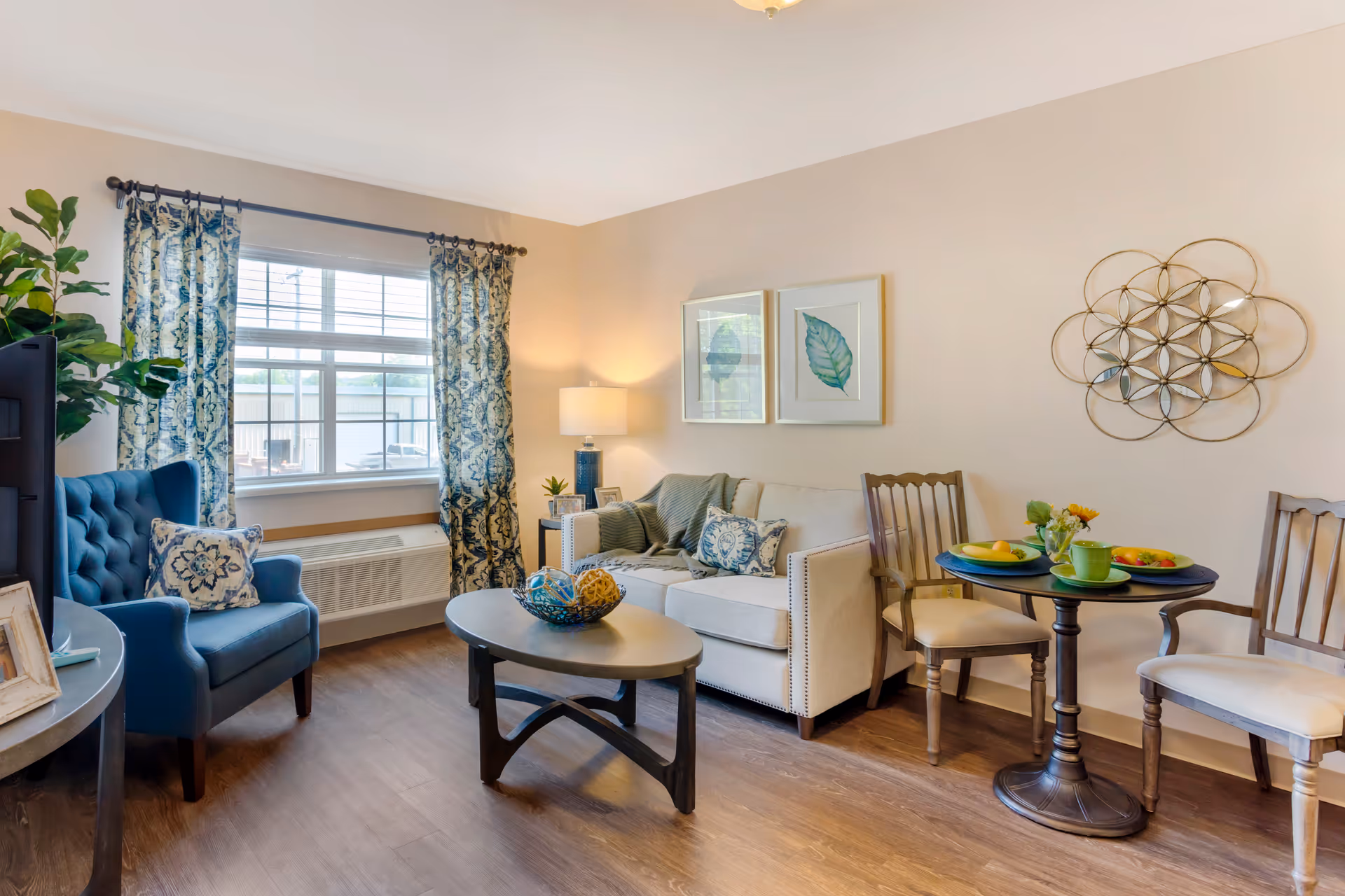 A cozy living room area with a blue armchair, a beige sofa with patterned cushions, a round coffee table with decorative items, and a small round dining table set with two chairs and plates of food. The room has a window with patterned curtains, a floor lamp, framed leaf artwork on the wall, and a decorative metal wall hanging.