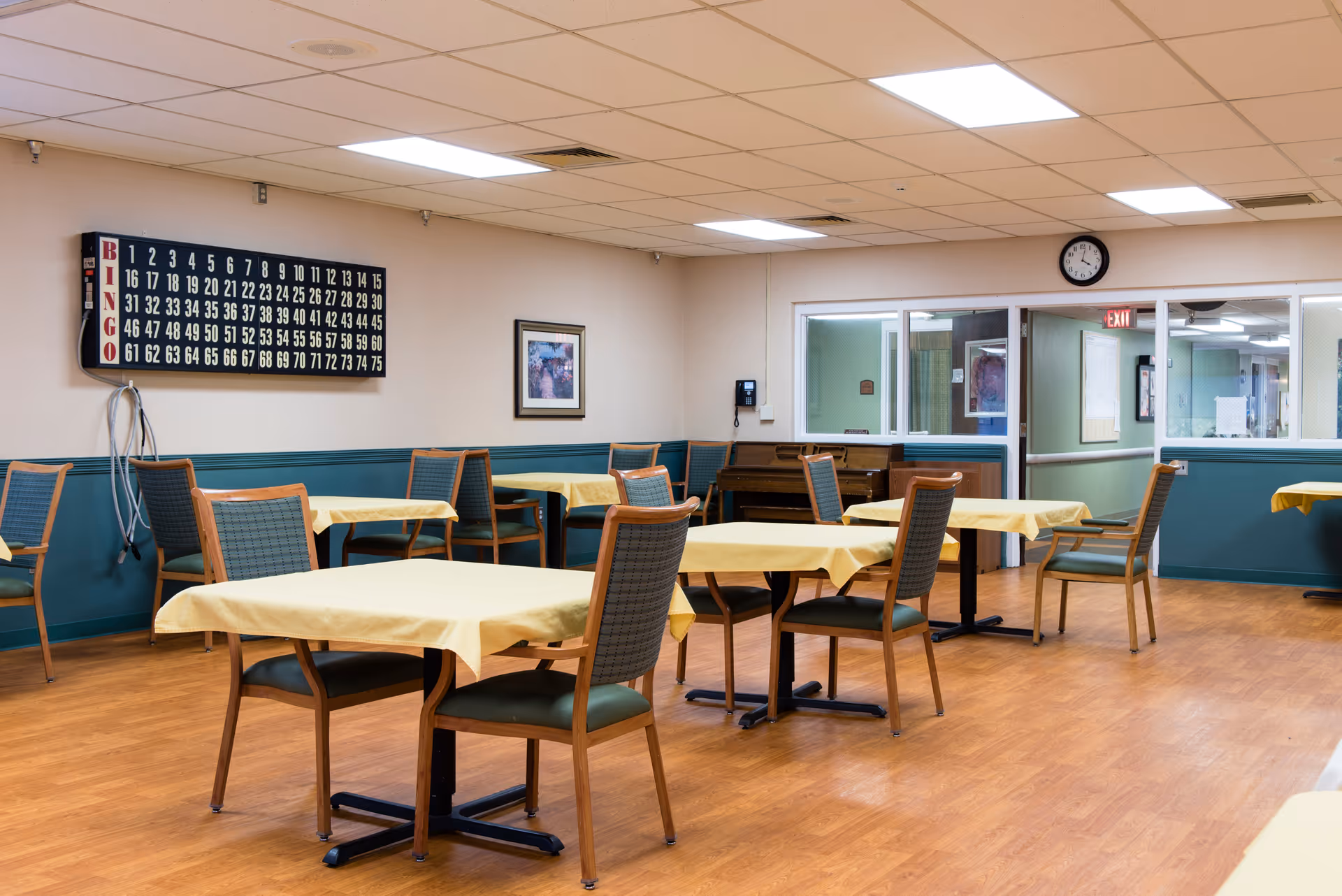 A well-lit room with several tables covered with yellow tablecloths and wooden chairs arranged around them. On the wall, there is a large bingo board with numbers and the word BINGO vertically displayed. The room has wood flooring, a clock on the wall, and a piano in the corner. There are windows and a door leading to another area.