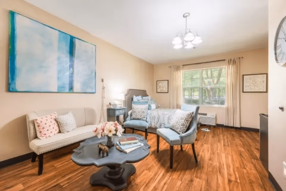 A cozy bedroom with wooden flooring featuring a bed with patterned bedding, two blue cushioned chairs with patterned pillows, a beige loveseat with decorative pillows, a dark wooden coffee table with books and flowers, a large window with beige curtains, and a wall clock.