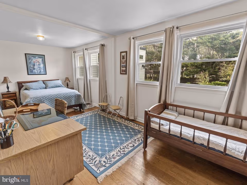 Sunlit bedroom with a double bed, wooden daybed, desk, area rug, and windows overlooking greenery.