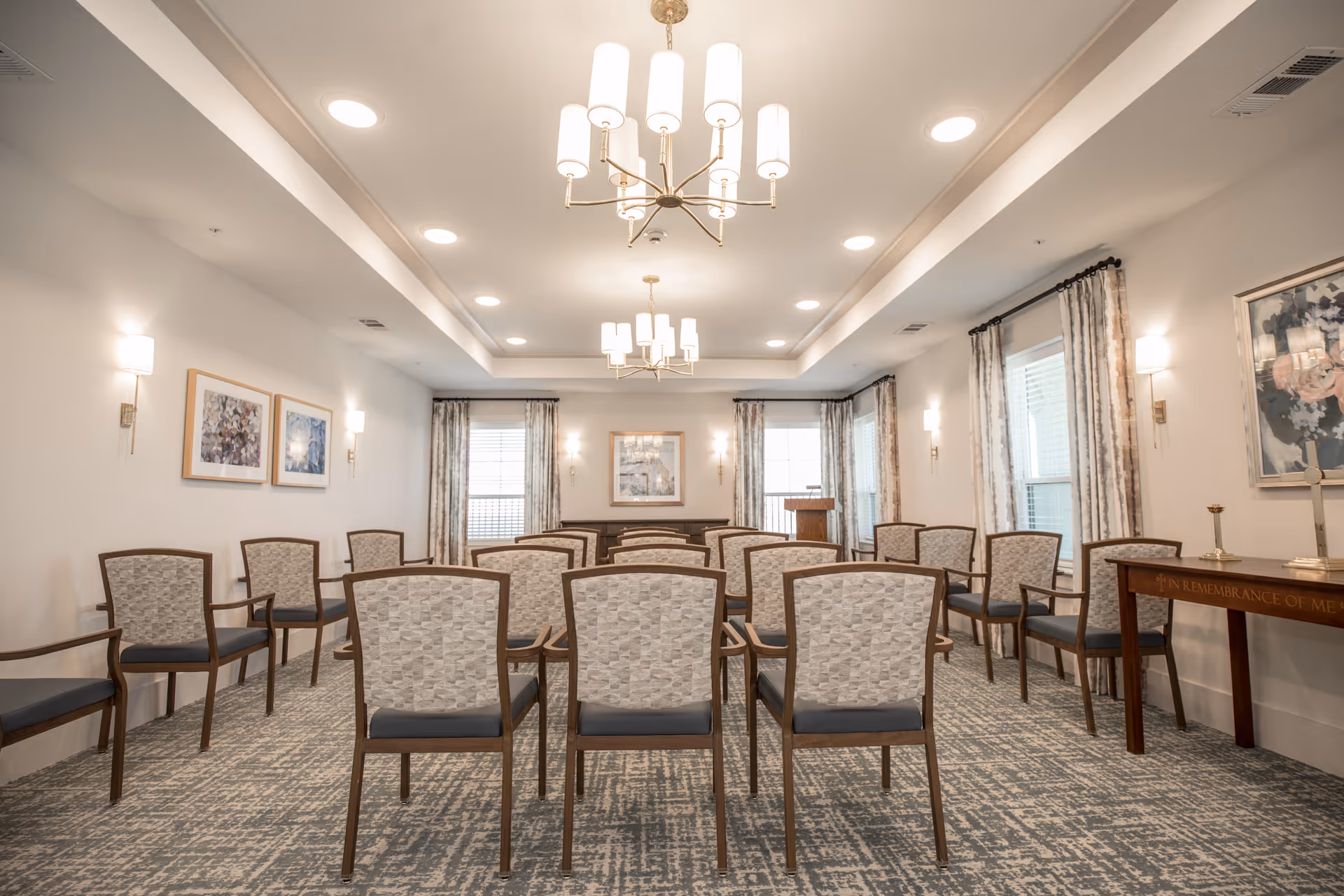 A well-lit room with rows of chairs arranged facing a podium and a table at the front. The room has patterned carpet, light-colored walls with framed artwork, large windows with curtains, and two chandeliers hanging from the ceiling.