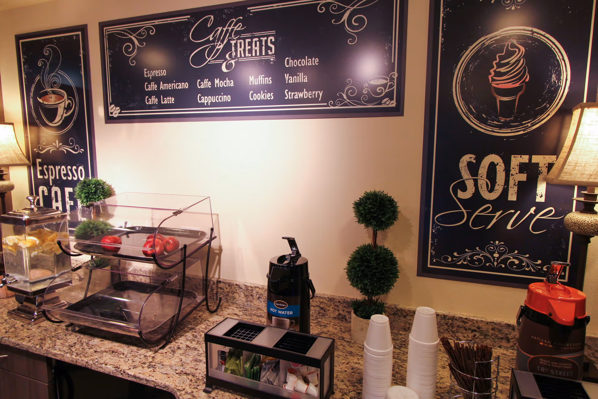 A coffee and treat station with a granite countertop featuring a water dispenser with lemon slices, a two-tiered clear display case with red apples, a hot water dispenser, disposable cups, and a container with stirrers. The wall behind has decorative signs reading 'Espresso Cafe', 'Caffe & Treats' listing coffee and treat options, and 'Soft Serve' with an ice cream cone illustration. Two small potted topiary plants and two table lamps are also visible.