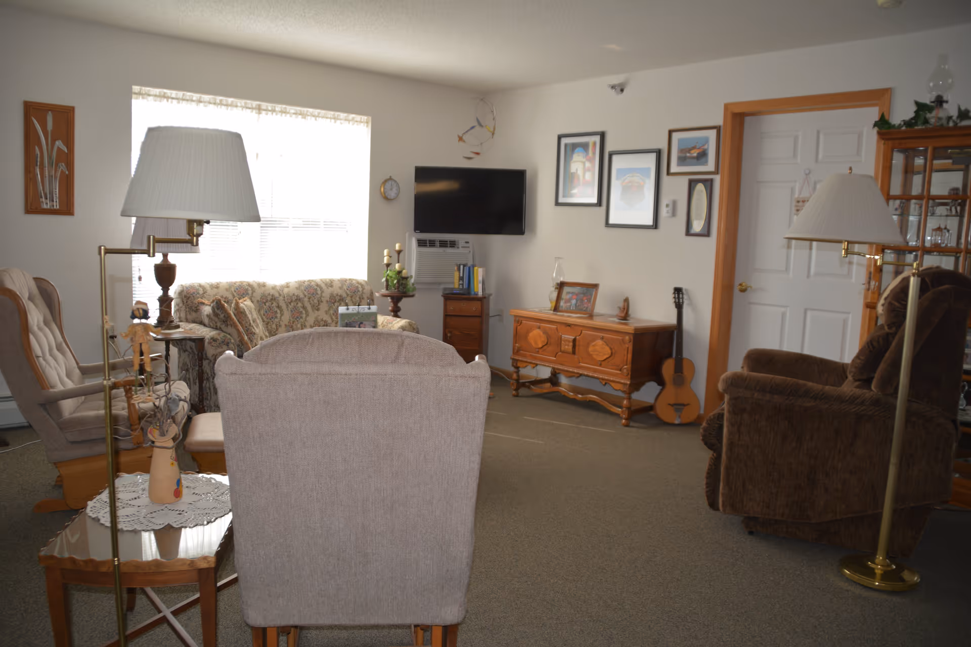 A cozy living room with a floral patterned sofa, two armchairs, a wooden side table with a lamp, a wooden cabinet with framed pictures and a guitar leaning against it, a wall-mounted TV, and several framed artworks on the walls.
