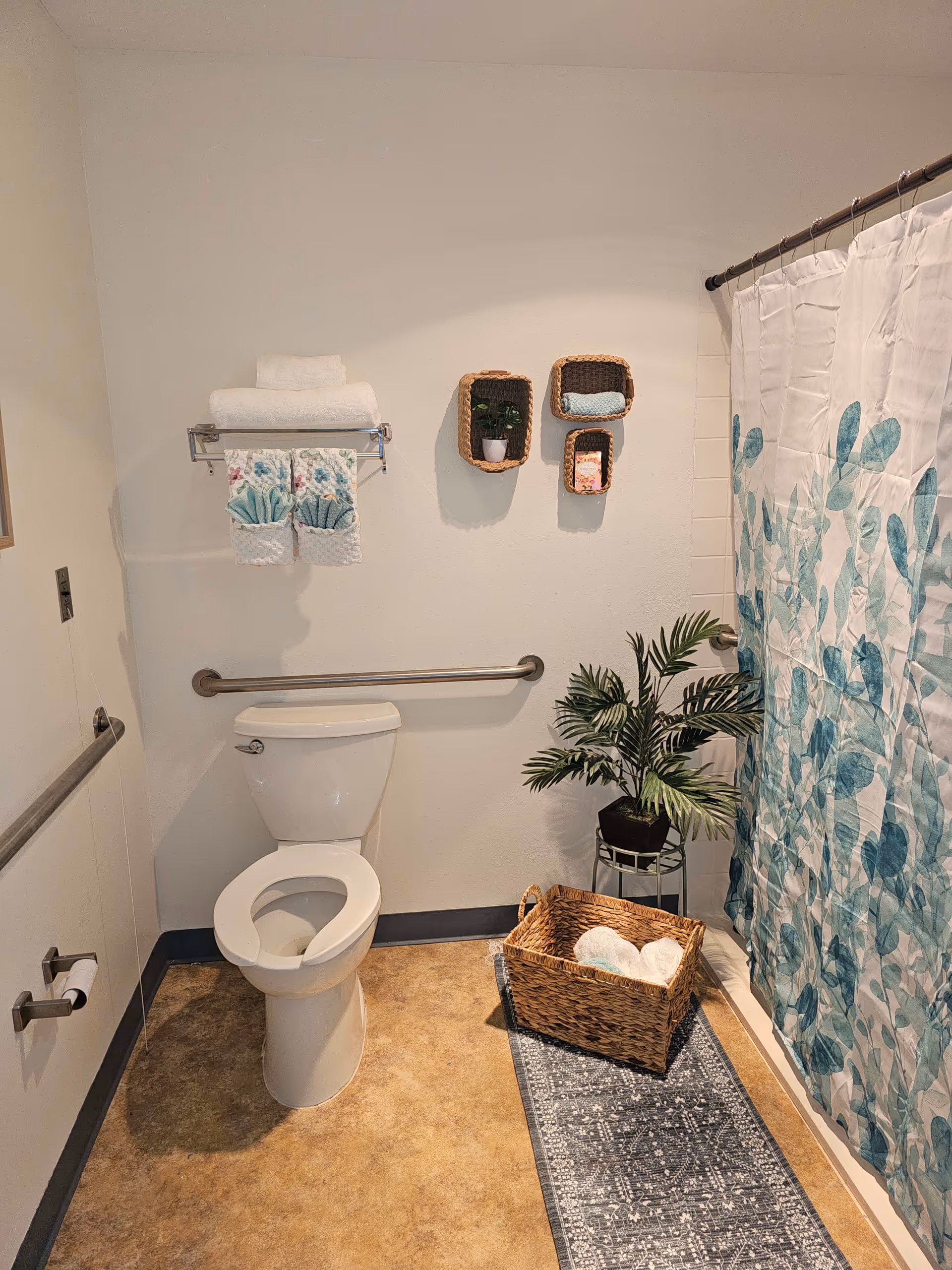 Accessible bathroom with a toilet, grab bars, towel rack, shower with a leaf-patterned curtain, decorative baskets and a potted plant.