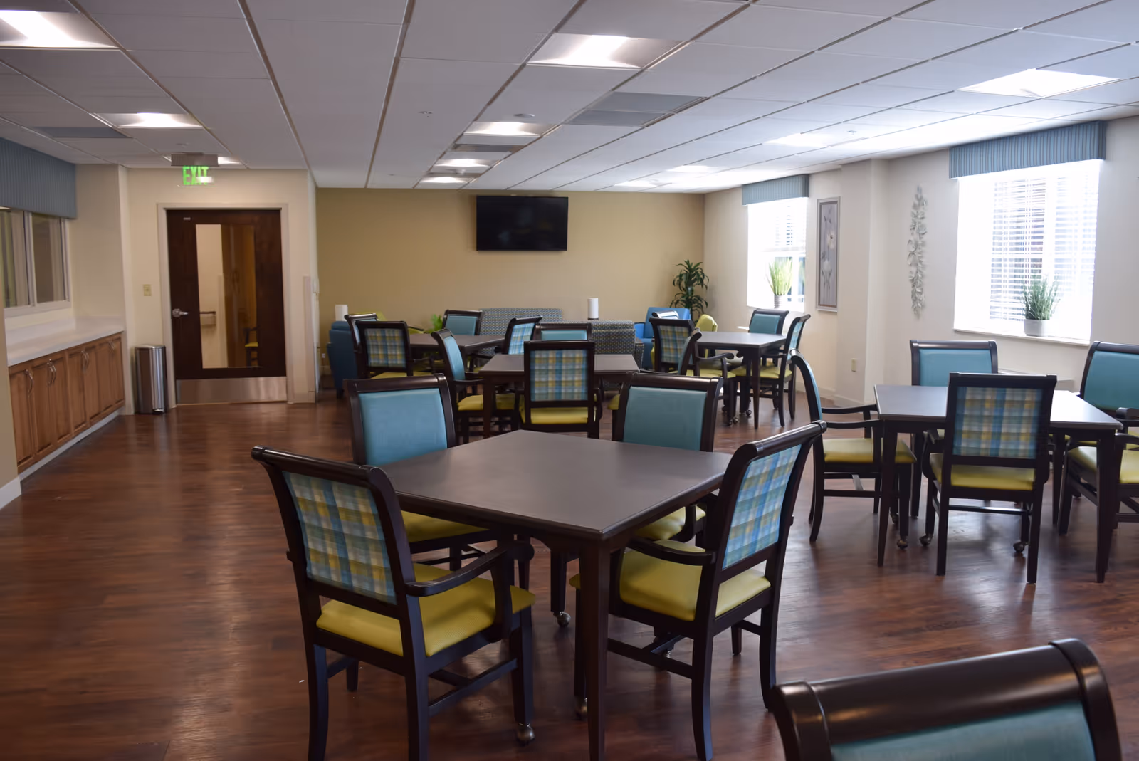 A bright and spacious dining room in a retirement home with several dark wood tables and chairs featuring blue and green plaid backs and yellow seats. The room has large windows with blinds, light-colored walls, wood flooring, and a wall-mounted TV. There are plants and framed artwork on the walls, and an exit door is visible at the back.