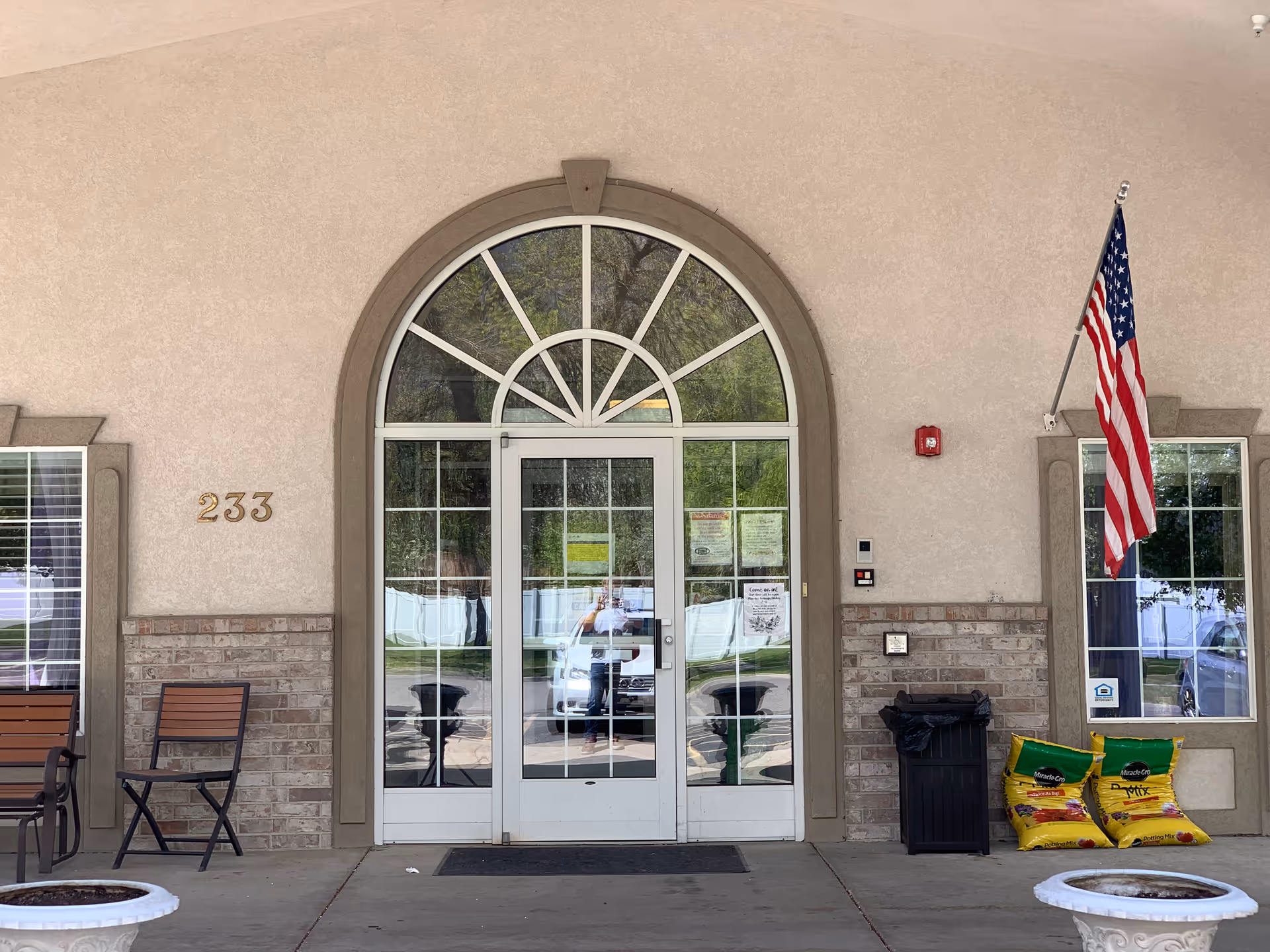 Front entrance of a senior living facility with an arched glass double door, the number 233 on the wall, an American flag, chairs, and bags of supplies nearby.