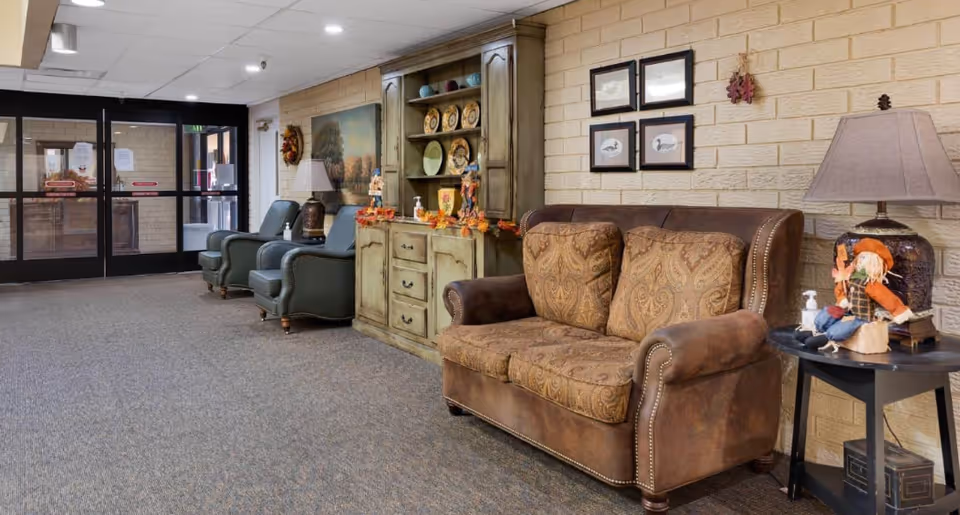 Seating area in a retirement facility lobby with a patterned leather sofa, two armchairs, a side table with a lamp and decorations, and a decorative cabinet against a brick wall.