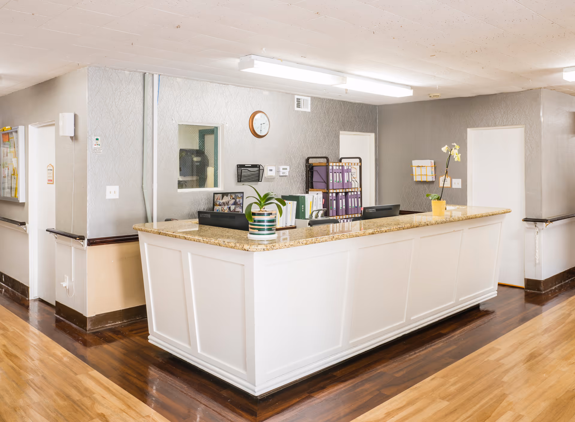 Reception desk area inside a nursing center with a granite countertop, two potted plants, office supplies, and file organizers. The walls are painted gray with a clock mounted above a small window. The floor is a combination of light and dark wood, and there are doors and handrails along the hallway.