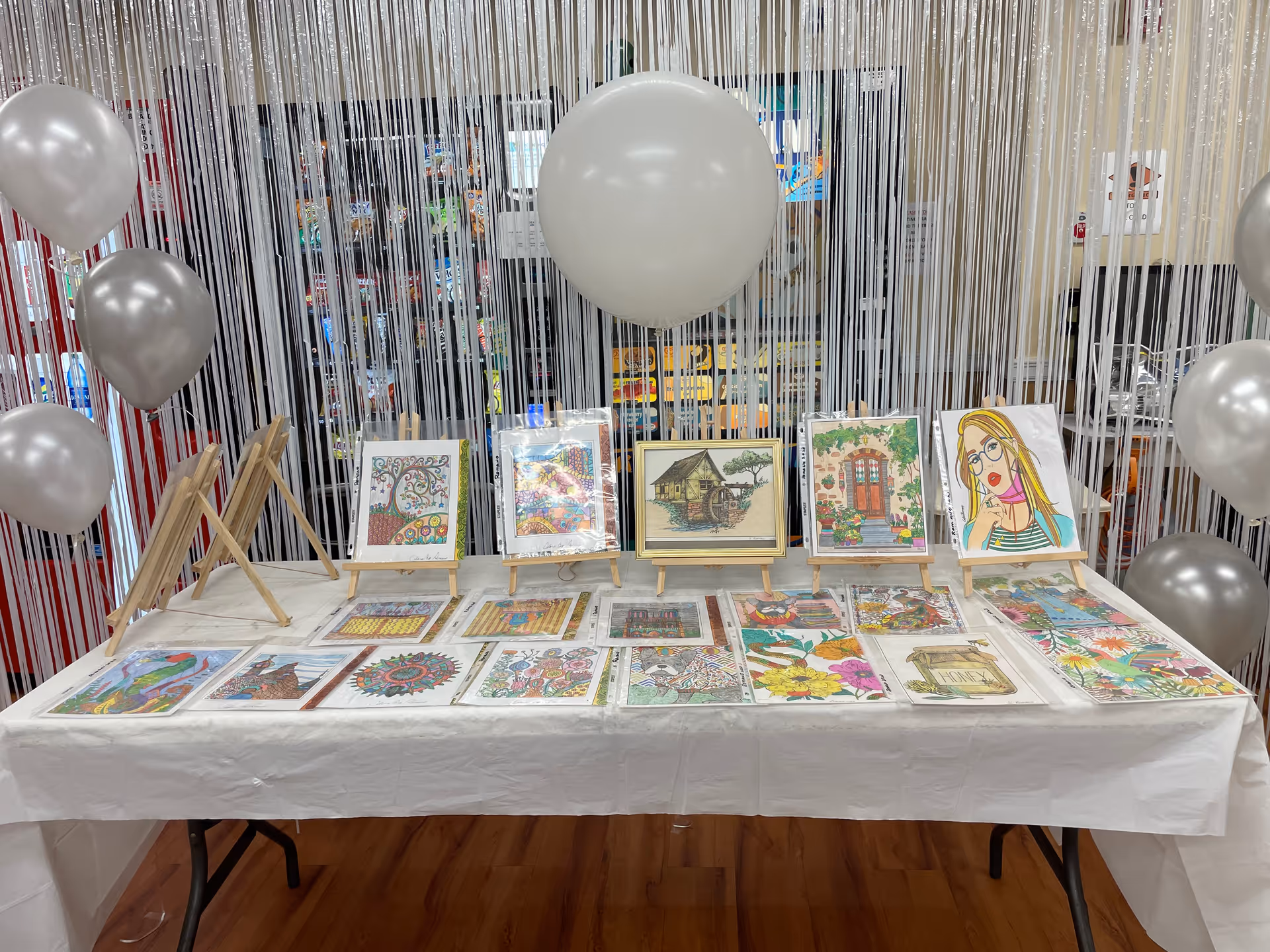 A display table covered with a white tablecloth showcasing various colorful artworks, including drawings of a tree, a house, flowers, and a woman with glasses. The artworks are arranged both on small wooden easels and laid flat on the table. Silver and white balloons are tied around the table, and a backdrop of vertical white streamers hangs behind it.