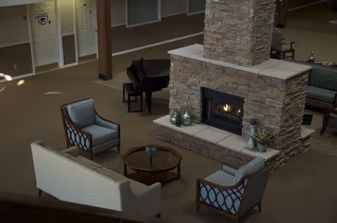 Cozy communal living area with a central stone fireplace, sofas and armchairs arranged around a glass coffee table and a piano in the background.