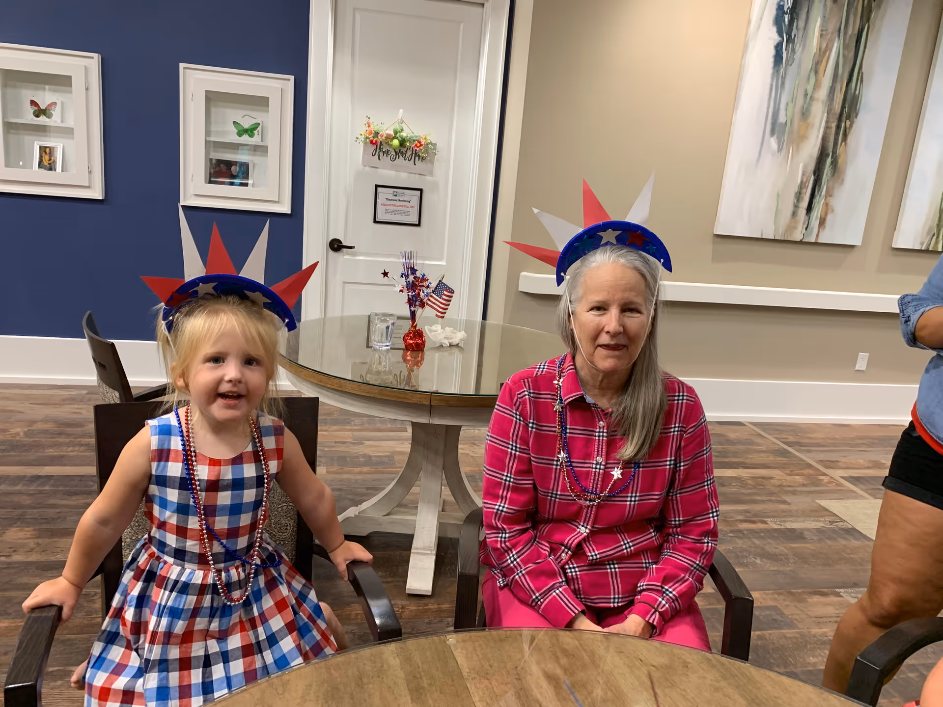 A young girl and an elderly woman sitting at a round wooden table in a room with wood flooring and neutral-colored walls. Both are wearing red, white, and blue star-spiked headbands and festive beaded necklaces. The girl is dressed in a red, white, and blue checkered dress, while the woman is wearing a red plaid shirt. Behind them is a glass-topped table with patriotic decorations and a door with a sign. Framed butterfly pictures and abstract paintings hang on the walls.