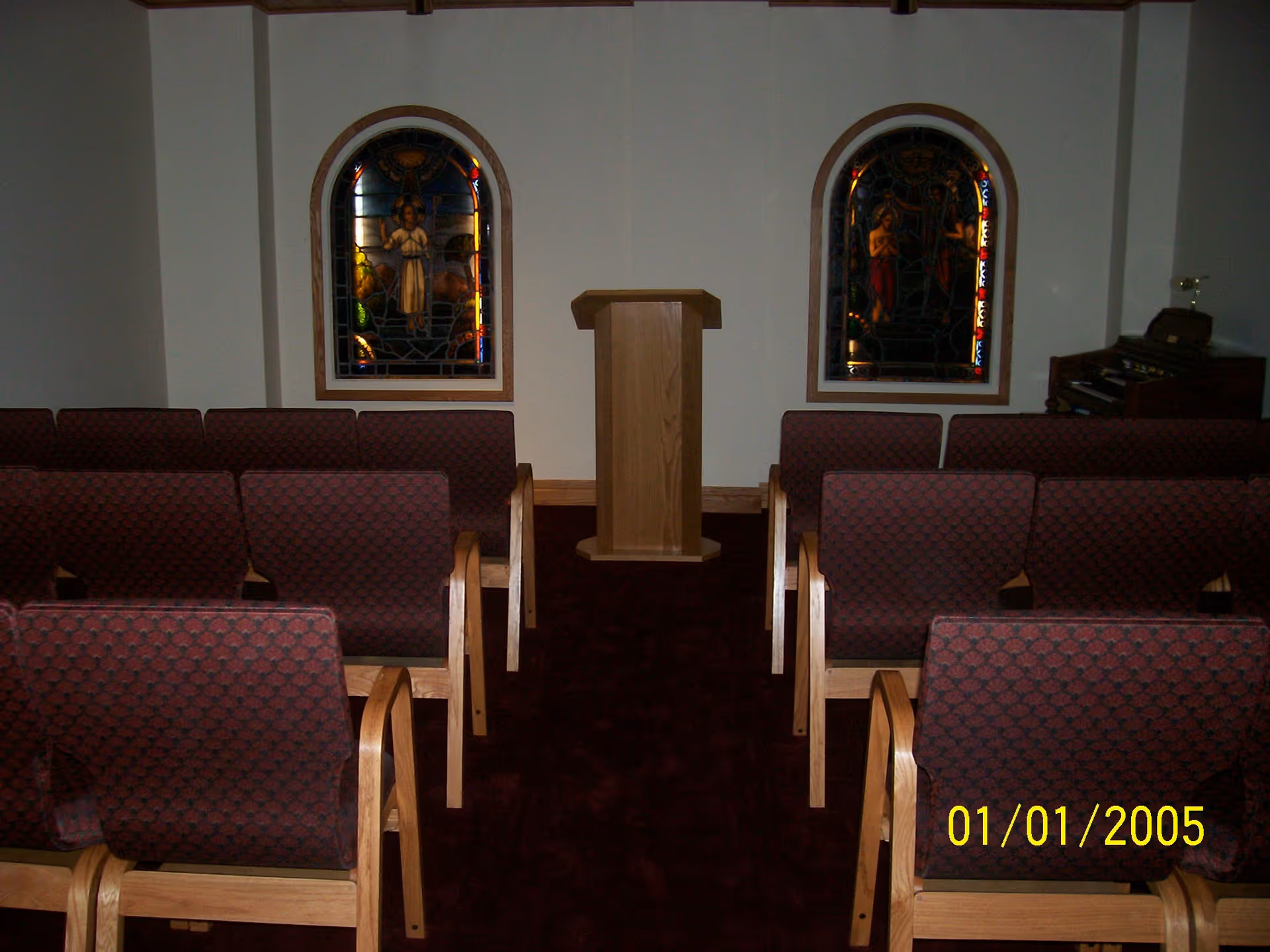Small chapel-like interior with rows of upholstered chairs facing a wooden lectern and two stained-glass windows.