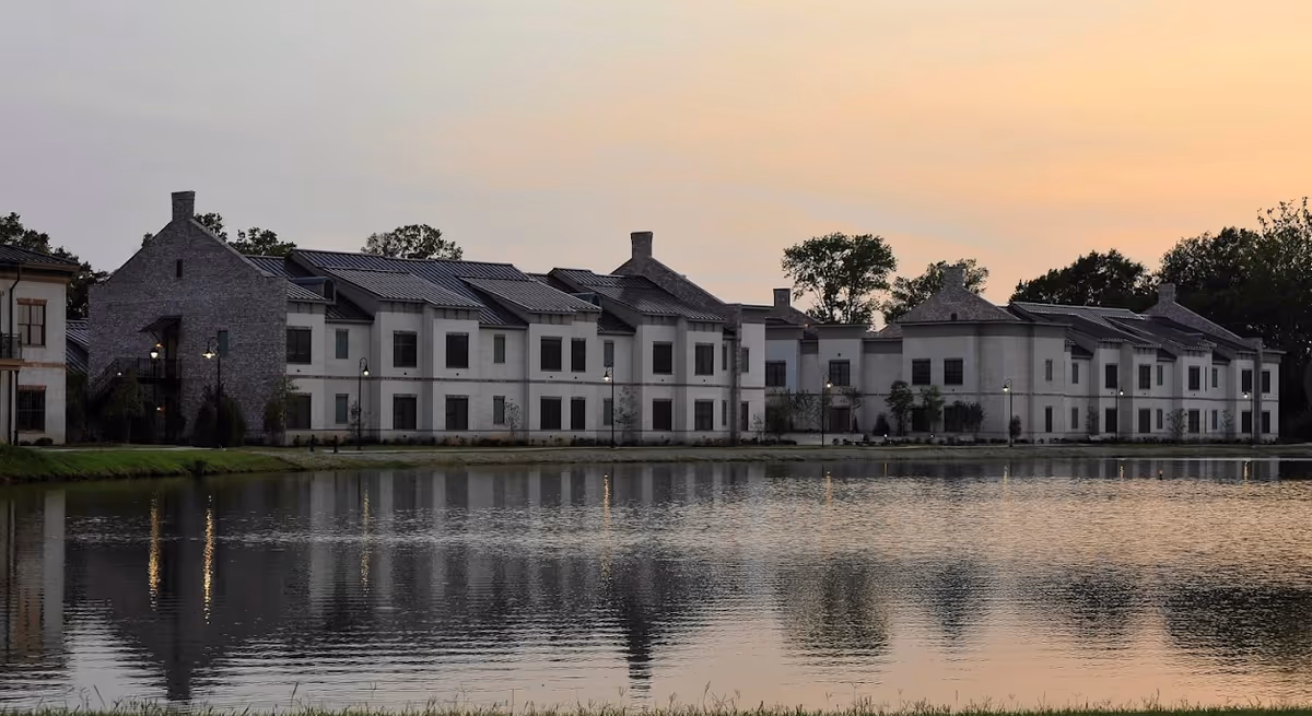Exterior view of The Blake at Township senior living facility at sunset, showing a large two-story building with multiple windows and chimneys, reflected in a calm pond in the foreground, with trees and soft evening sky in the background.