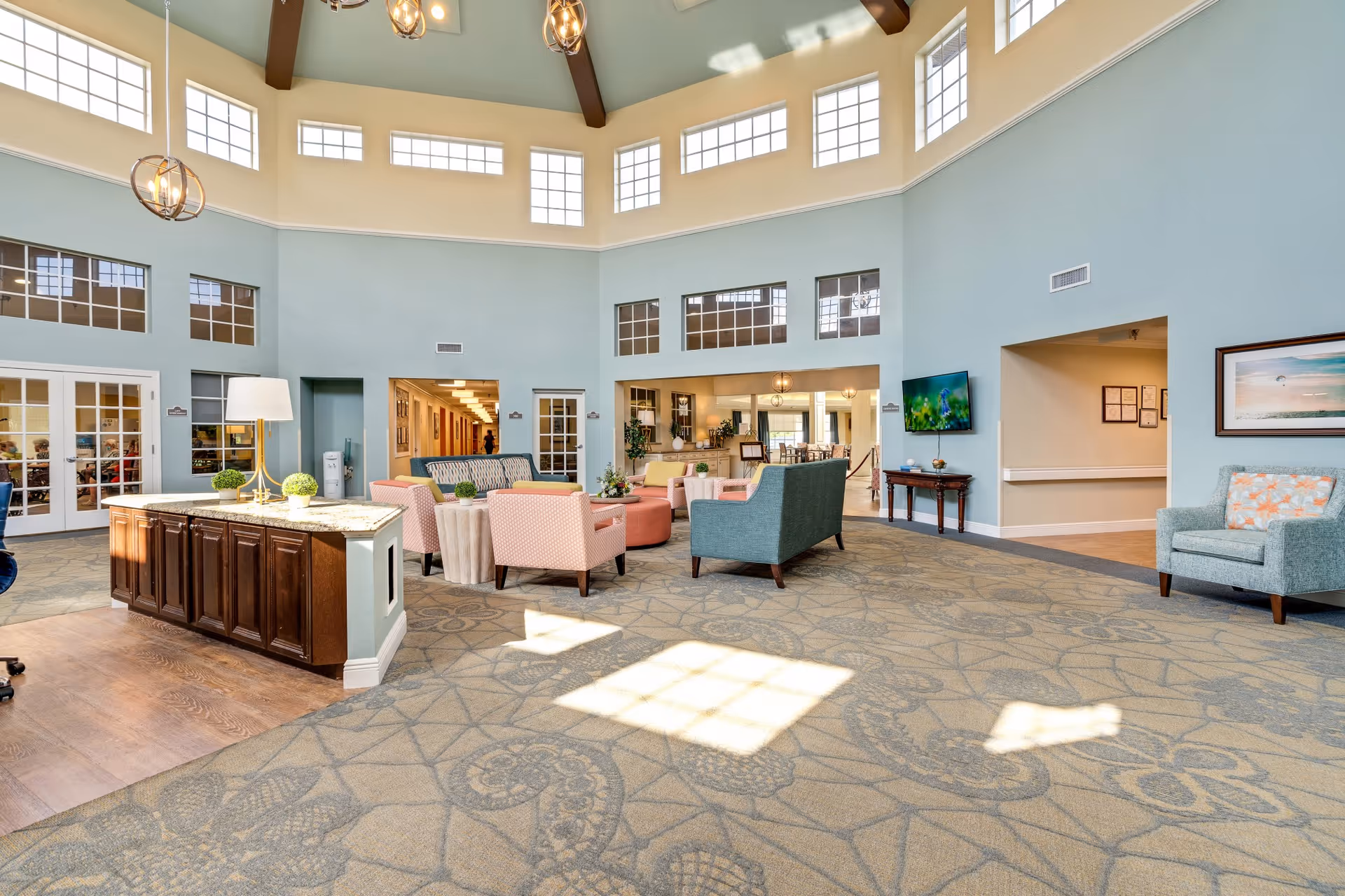 A spacious and well-lit common area in a senior living facility with high ceilings and multiple windows. The room features comfortable seating including armchairs and sofas in pastel colors, a wooden cabinet with a lamp and decorative plants, a wall-mounted TV, and framed artwork. The carpet has a patterned design, and there are doorways leading to other rooms.