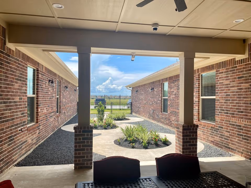 View from a covered patio area looking out onto a landscaped courtyard with circular planting beds and brick walls on either side. The sky is blue with some clouds, and there is a metal fence at the far end of the courtyard.