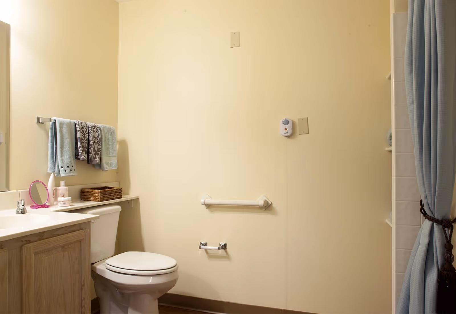 A bathroom with a toilet, a vanity with a sink, a mirror, and a towel rack holding three towels. There is a grab bar on the wall next to the toilet and a shower curtain partially visible on the right side.