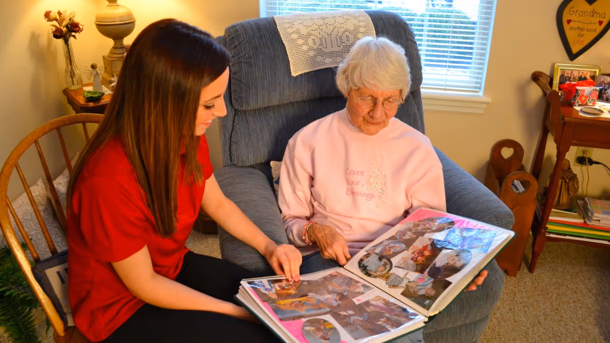 An elderly woman with white hair wearing a pink sweatshirt sits in a blue armchair looking at a photo album with a younger woman with long brown hair wearing a red shirt. They are in a cozy, well-lit living room with a wooden chair, a side table with a lamp and flowers, and a window with blinds in the background.
