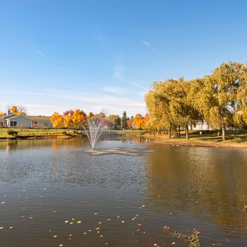 A peaceful pond with a water fountain in the center, surrounded by trees with autumn foliage and residential buildings in the background under a clear blue sky.