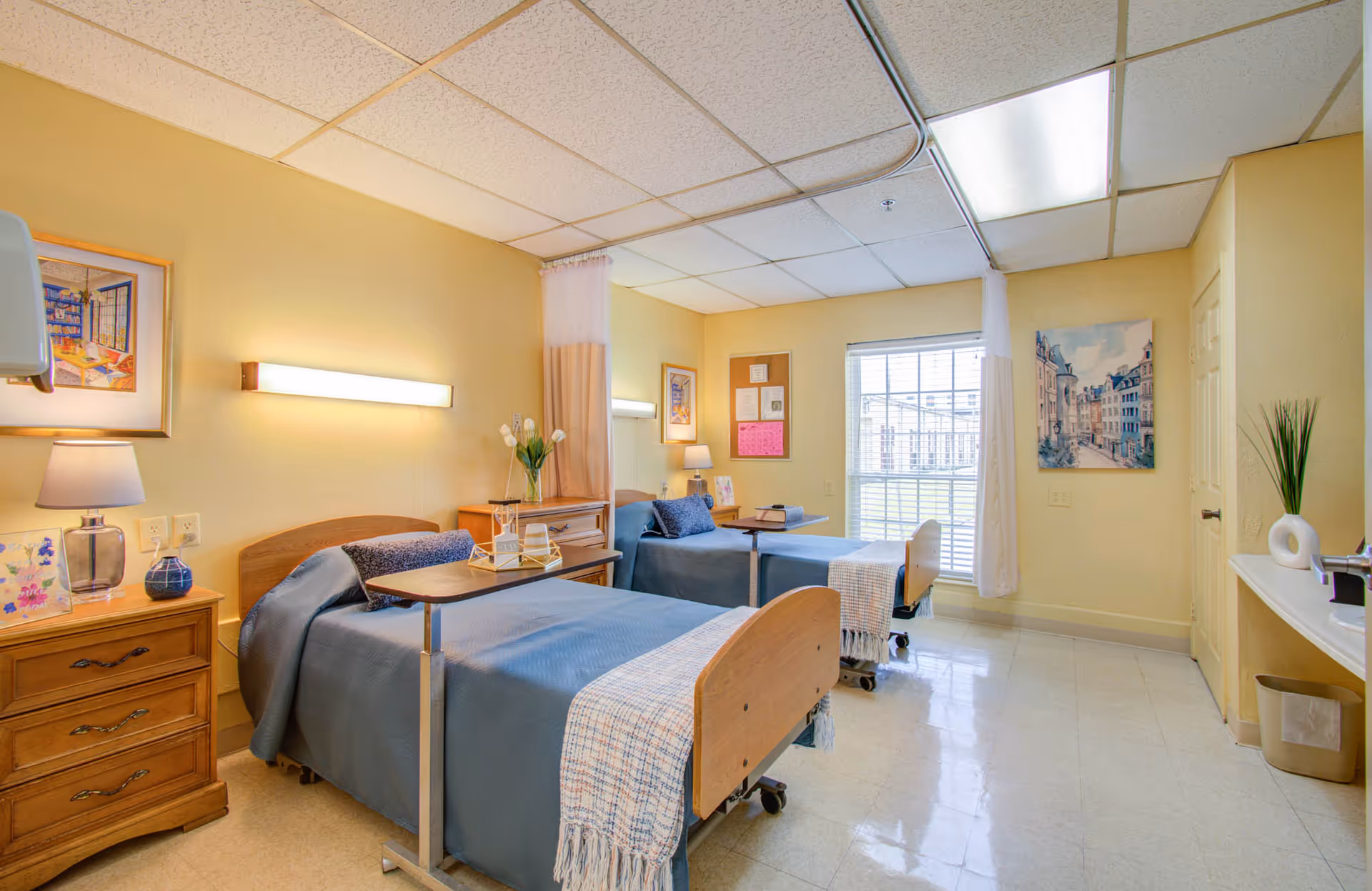 Bright shared nursing facility bedroom with two hospital-style beds, overbed tables, nightstands, and a window.