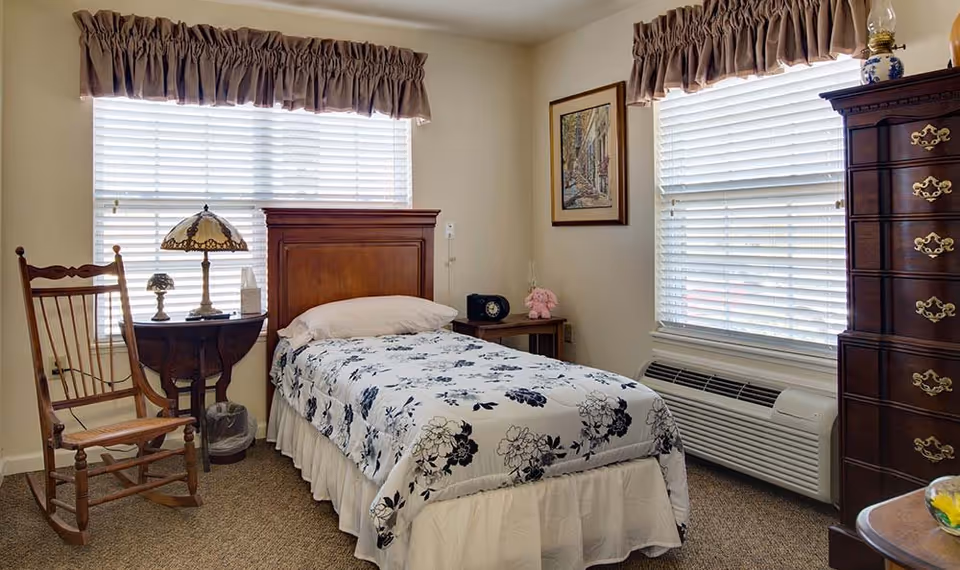 A cozy bedroom with a single bed covered in a white and black floral bedspread. The room features two windows with blinds and valances, a wooden rocking chair, a small round table with a decorative lamp and tissue box, a wooden nightstand with a clock and a pink stuffed animal, and a tall wooden dresser with brass handles.