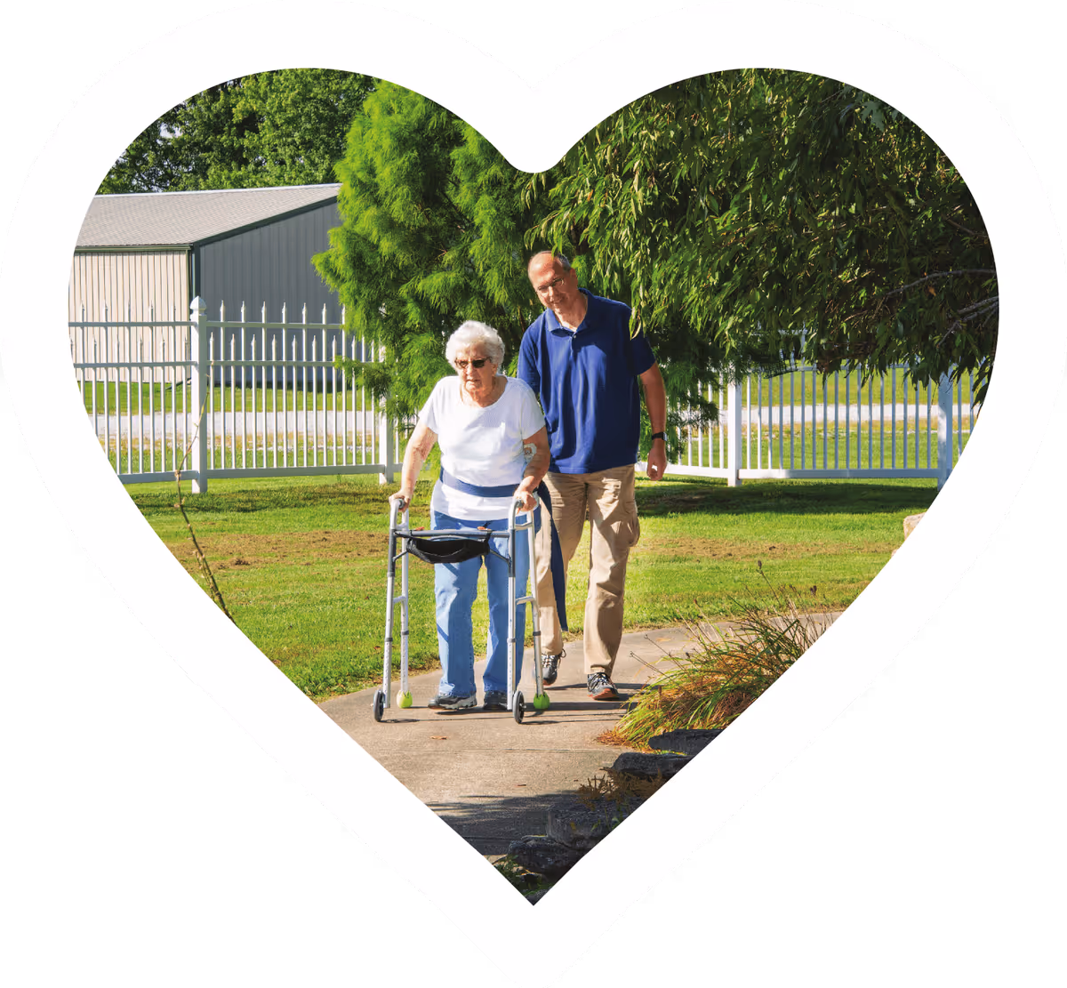 An elderly woman using a walker is walking on a paved path outdoors, accompanied by a man walking beside her. They are in a green grassy area with trees and a white fence in the background. The image is framed within a white heart shape.