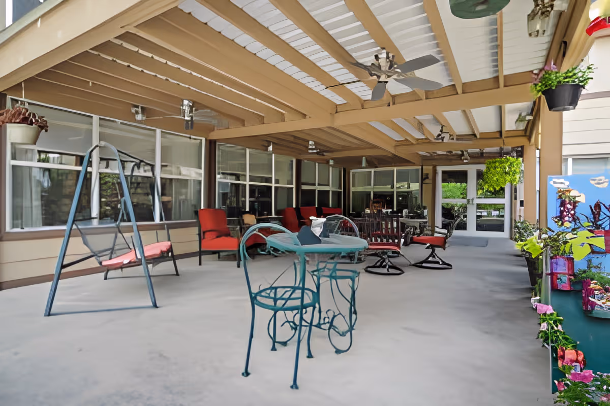 Covered outdoor patio area with a variety of seating including red cushioned chairs, a metal swing with red cushions, and a small metal table with two chairs. The patio has a beige and white ceiling with ceiling fans and hanging plants. Large windows and a door lead into the building.